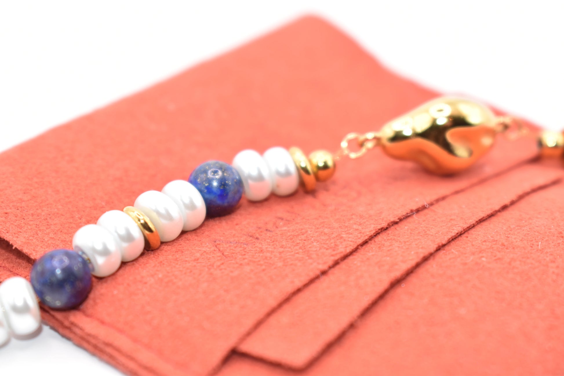 Close-up of bracelet with white pearls, blue lapis beads, and gold accents on coral background