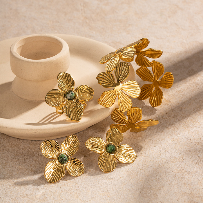 A collection of golden flower-shaped earrings and pins, beautifully textured to resemble petals, each adorned with a green gemstone at the center, presented on a textured neutral-colored backdrop.
