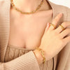  a woman wearing a luxurious gold link necklace paired with matching rings and a bracelet. The jewelry is prominently featured against her beige sweater, highlighting its intricate links and sleek design. The soft focus on her hands subtly draws attention to the rings, while the smooth texture of the fabric provides a neutral backdrop that enhances the jewelry's shine.