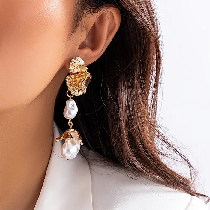 The earring features a detailed gold leaf at the top, closely resembling natural leaf texture with its ridges and veins highlighted in gold. Suspended from this leaf are two pearls, each unique in shape and lustrous in appearance, followed by a delicate gold pendant shaped like a teardrop. The woman is wearing a white blouse, providing a neutral background that accentuates the warm tones of the gold and the soft glow of the pearls. 