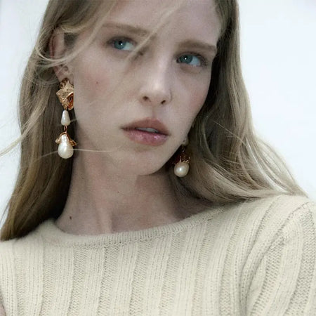 The earrings are designed with gold leaf motifs and dangle elegantly from her ears, each adorned with a baroque pearl and a smaller gold pendant. She is dressed in a soft, cream-colored sweater, complementing the warm tones of the jewelry. Her hair is light blonde, loosely styled, and her expression is contemplative