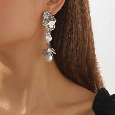 The image features a close-up view of a woman wearing Aimee Leaf Silver Pendant Earrings. The earrings are designed with a silver leaf at the top, artistically detailed to capture the natural texture of a leaf. Below the leaf, two baroque pearls are suspended, adding a touch of elegance. The model has her hair pulled back to better showcase the earrings against her bare neck, enhancing the visual appeal of the jewelry against a simple black top.