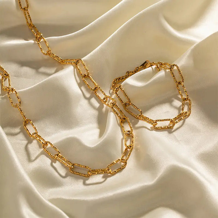  a gold paperclip chain bracelet and necklace gracefully placed on a smooth, lustrous satin fabric, highlighting their elegant design and luxurious appearance. The golden hue of the jewelry contrasts beautifully against the creamy white background, emphasizing the intricate link patterns and the overall sophistication of the pieces.