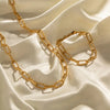 a luxurious gold paperclip chain necklace and matching bracelet elegantly arranged on a smooth, cream-colored satin fabric. The golden jewelry pieces have detailed texturing, adding an exquisite and refined look that catches the eye with their intricate design. The soft folds of the satin background provide a rich and opulent setting that highlights the luster of the gold, showcasing the jewelry's sophisticated appeal.