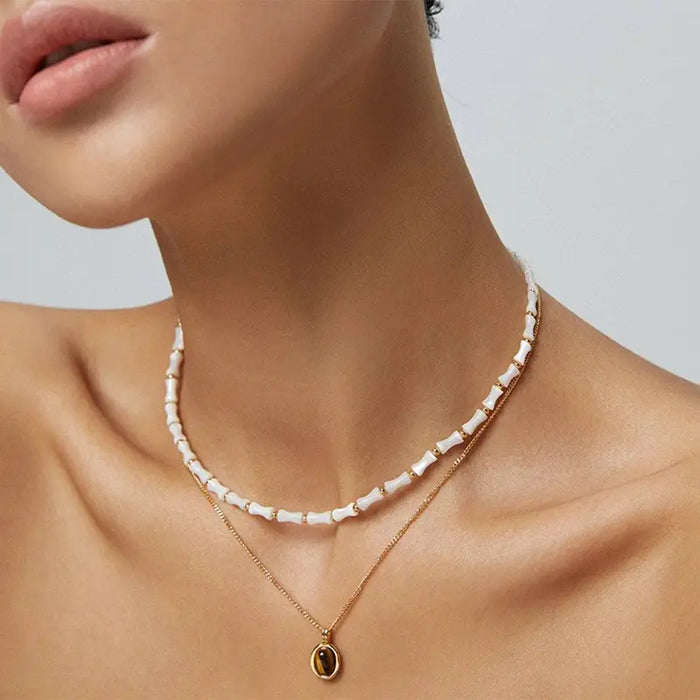 Model wearing a layered Anais & Aimee Tiger's Eye Pendant Necklace featuring a chic white beaded choker and a delicate gold chain with an oval tiger`s eye gemstone pendant.