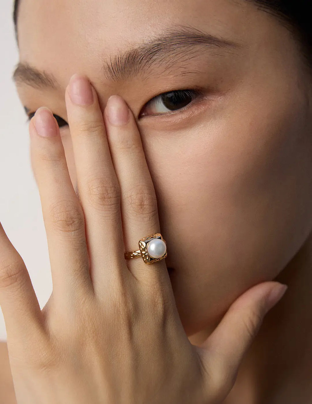 Close-up of a hand wearing a gold ring with a pearl on a neutral background