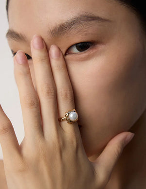 Close-up of a hand wearing a gold ring with a pearl on a neutral background