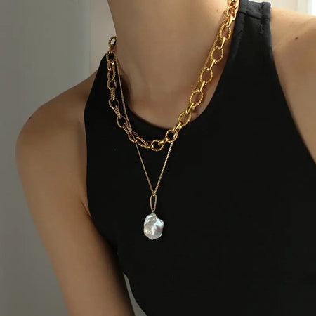 The large, lustrous pearl hangs from a delicate gold chain, which is paired with a thicker, more ornate gold link chain worn closer to the neck. This layered jewelry style creates a sophisticated and contemporary look. The backdrop of her black outfit provides a striking contrast that accentuates the golden hues of the chains and the iridescent quality of the pearl. This ensemble exemplifies a chic and modern approach