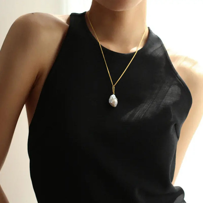 The large, irregularly shaped pearl provides a lustrous focal point against the backdrop of her sheer black top, which offers a subtle contrast that highlights the pearl's natural beauty and radiant shine. The minimalistic style of the necklace makes it a versatile accessory that complements a chic and modern wardrobe, perfect for both casual and formal occasions. The overall presentation emphasizes simplicity and elegance, making the necklace a timeless piece of jewelry.