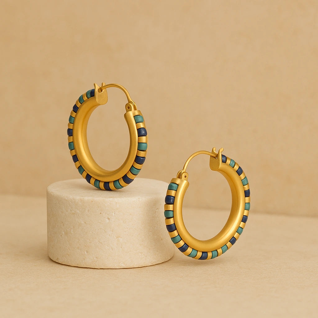Gold-plated hoop earrings with turquoise, lapis, and green gemstone beads displayed on a round neutral pedestal.

