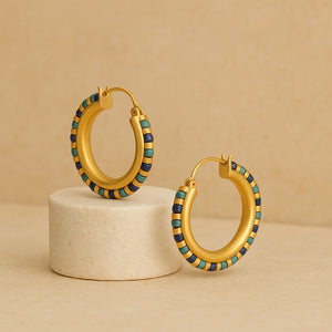 Gold-plated hoop earrings with turquoise, lapis, and green gemstone beads displayed on a round neutral pedestal.

