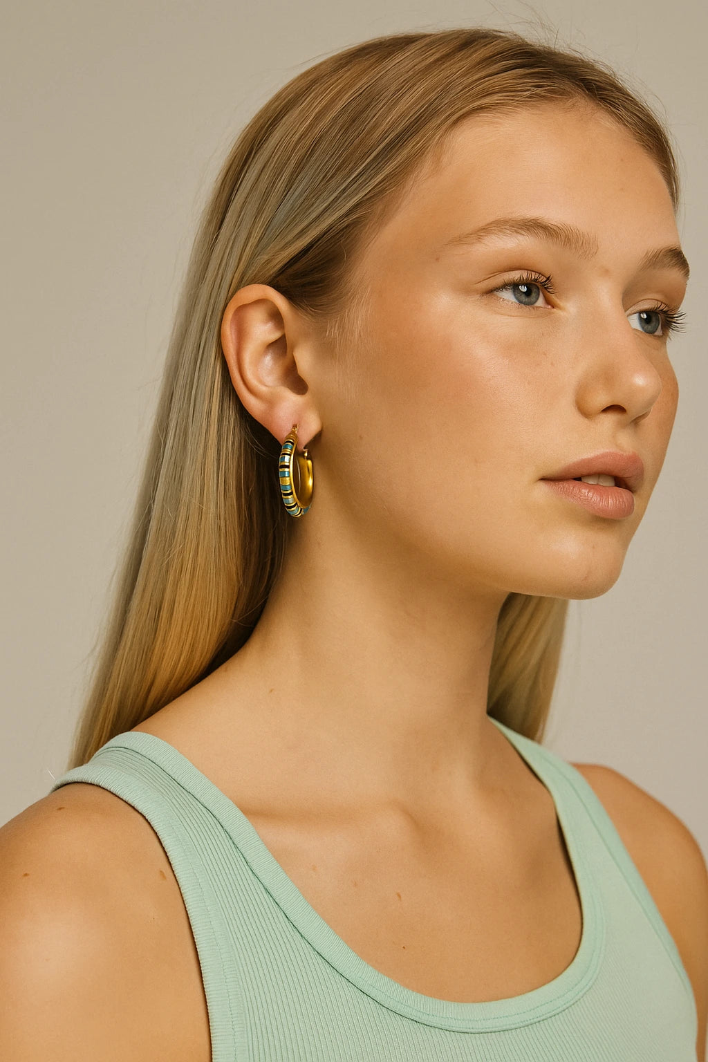Close-up of a model wearing a single gold beaded turquoise hoop earring with natural makeup and mint green top.


