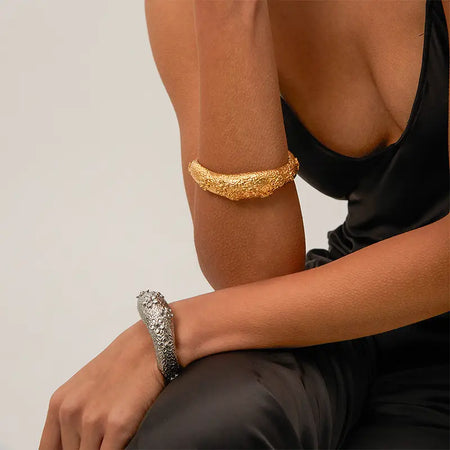 Model wearing the Blooming Flower Textured Cuff in gold on her upper arm and a silver version on her wrist, showcasing the intricate floral motifs and textured finish.