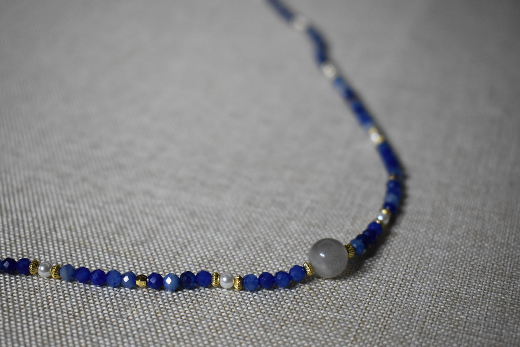 Close-up of Camile Turquoise Lapis Lazuli Pendant Necklace, highlighting the intricate blue lapis lazuli beads with gold detailing, resting on a grey fabric.