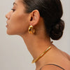 a profile view of a woman wearing striking large gold hoop earrings that curl close to her ears. These contemporary C-curve earrings have a bold and sculptural design, emphasizing a minimalist yet elegant aesthetic. The earrings make a significant statement and are likely to be a focal point of any ensemble,