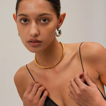  a close-up of a woman wearing unique and stylish jewelry. She has large, silver hoop earrings that have a reflective, polished finish, and a minimalist gold choker around her neck, enhancing her elegant look. Her bare shoulders and subtle makeup, combined with her direct gaze, give a sophisticated and contemporary feel to the image, focusing on the contrast and interaction of the cool-toned silver with the warm gold.