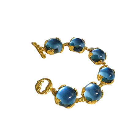 an elegant bracelet adorned with large, deep blue spherical gems set in ornate golden mounts that evoke a sense of regal opulence. Each gem is encircled with detailed gold filigree, enhancing the rich blue hue of the stones, which likely represent lapis lazuli or a similar semi-precious stone. The bracelet clasps with a delicate, yet intricately designed gold link that complements the overall aesthetic. 