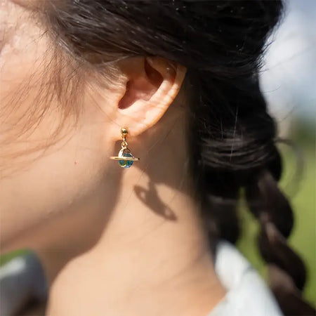 Close-up of a woman's ear wearing the Dainty Jupiter Earrings, featuring a blue gem encircled by a golden ring, simulating a miniaturized planet orbiting in space. The background is softly blurred, emphasizing the intricate and elegant design of the earrings