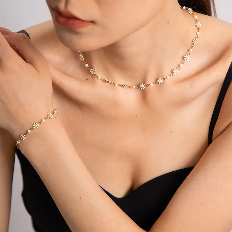 a woman elegantly wearing a dainty jewelry set consisting of a necklace and a bracelet. Both pieces are adorned with small white daisy-like flowers, each centered with a golden dot, spaced evenly along a delicate gold chain. This subtle and charming design adds a touch of springtime grace to her outfit, emphasizing a minimalist yet sophisticated aesthetic. 