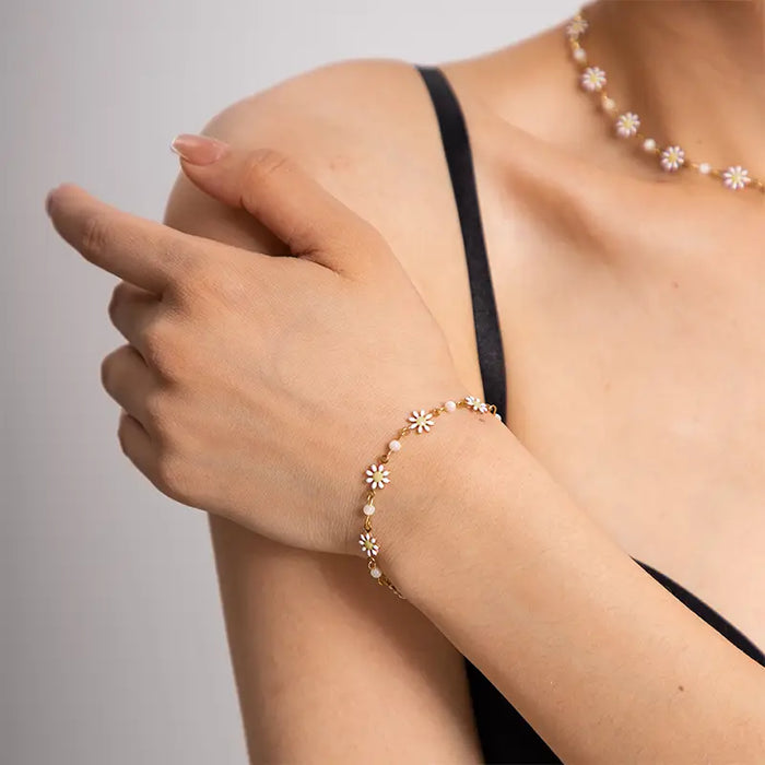 a close-up view of a woman's wrist adorned with a delicate bracelet from a jewelry set. The bracelet consists of small, white daisy-like flowers with golden centers, evenly spaced on a fine gold chain. This elegant piece complements her minimalist black attire, highlighting a subtle yet sophisticated style. The jewelry's design brings a touch of floral charm, making it a perfect accessory for both casual and formal occasions.