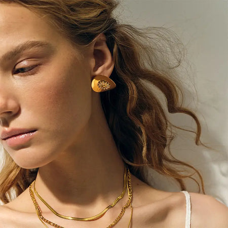 elegantly showcases a young woman adorned with golden dome earrings that feature a charming daisy design at their center. The smooth, polished gold of the earrings complements her soft, sun-kissed complexion and wavy, tousled hair, enhancing her natural beauty. The earrings, with their playful floral motif, bring a touch of whimsy to her sophisticated appearance, creating a balance between modern style and classic elegance. 