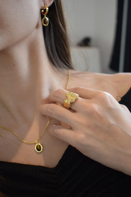 Close-up of a model's hand adorned with the Erosia Organic Gold Statement Ring, Anaïs & Aimee’s Tiger’s Eye Pendant Necklace, and minimalist gold drop earrings.