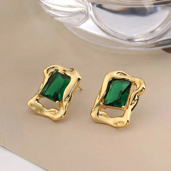 Evergreen Emerald Stud Earrings featuring vibrant green emerald-cut gemstones set in polished gold-tone metal with a unique, irregular frame design. These high trending earrings add a touch of timeless elegance and sophistication, perfect for making a stylish statement.