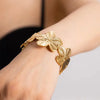Close-up of a model's wrist wearing a gold cuff bracelet adorned with large, detailed floral motifs, showcasing an elegant and polished finish.