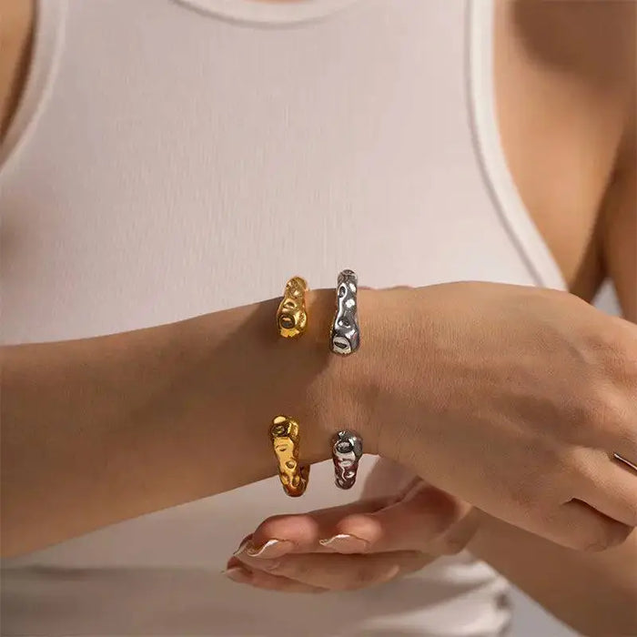 Woman wearing the Hammer Gold Bracelet and a matching silver bracelet, both featuring a unique hammered texture with raised, organic patterns. These high trending accessories are perfect for making bold and sophisticated fashion statements, adding a touch of elegance to any ensemble. She is dressed in a sleeveless white top, highlighting the bracelets' stylish appeal.