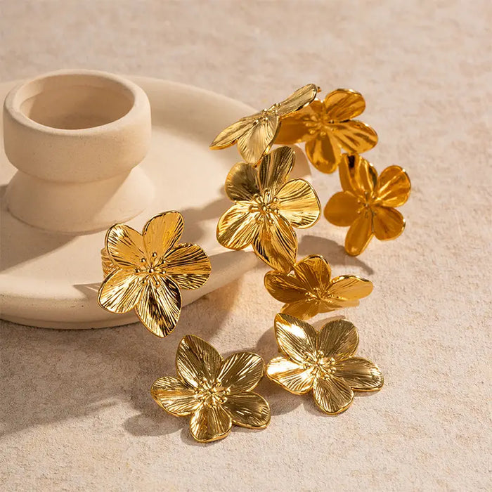 Set of gold hibiscus flower earrings with detailed petals, displayed on a beige surface with a ceramic holder, showcasing their elegant and polished design.