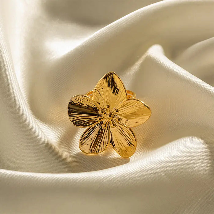 Gold ring featuring a detailed hibiscus flower design with polished petals, displayed on a soft, champagne-colored fabric.