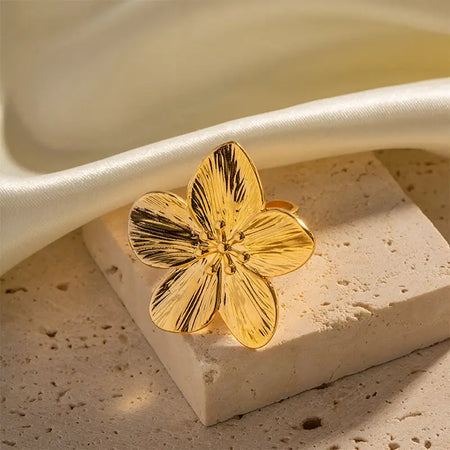 Gold ring featuring a detailed hibiscus flower design with polished petals, displayed on a textured stone surface with a soft, champagne-colored fabric in the background.