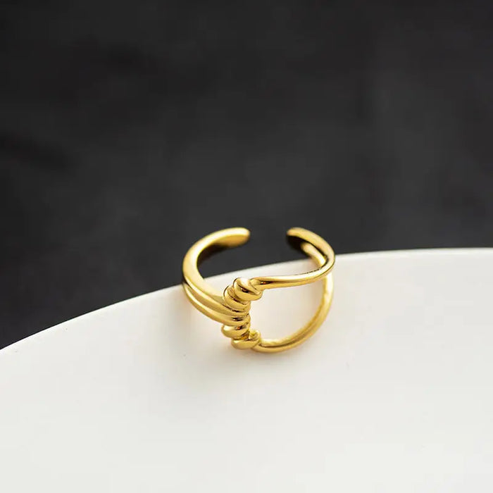 Knot Gold Ring featuring a unique open-ended design with a central knot detail, crafted in polished gold-tone metal. This high trending accessory adds a touch of modern elegance and sophistication, perfect for any occasion, displayed on a contrasting black and white background.