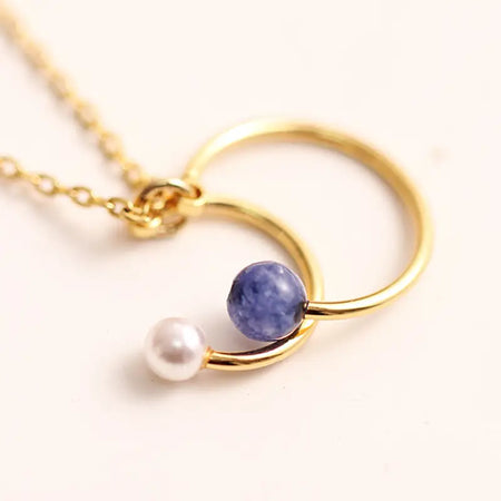 Close-up of the Anais & Aimee Lapis Lazuli Drop Necklace, featuring a gold chain with interlocking circular pendants, adorned with a deep blue lapis lazuli stone and a lustrous white pearl. Perfect for adding a touch of elegance and sophistication to any outfit.