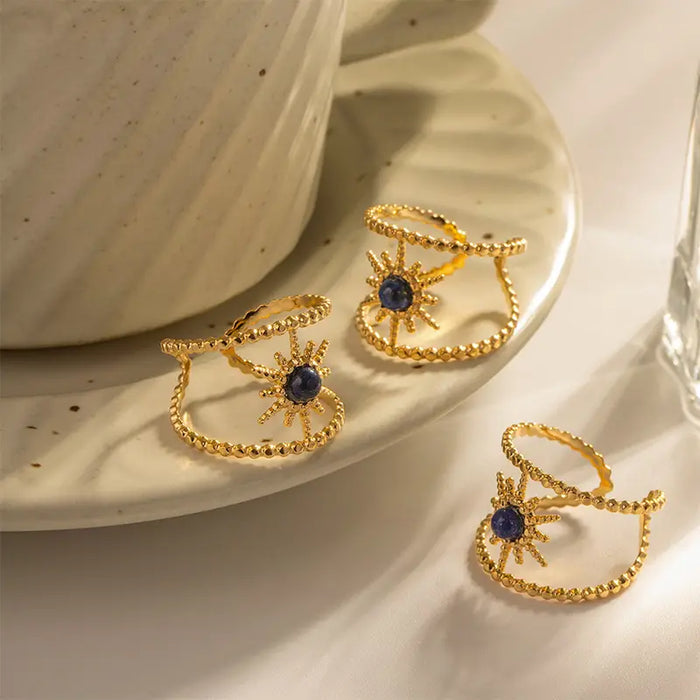 Three elegant gold bracelets with lapis lazuli stones set in a sunburst design, displayed on a marbled dish. Each bracelet features a radiant blue center surrounded by golden rays, with a twisted rope-style band, creatively arranged on a smooth, cream-colored ceramic surface.