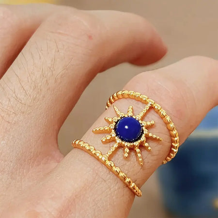 a close-up view of a lapis lazuli sunbeam bracelet worn on a person's finger. The bracelet features a prominent deep blue lapis lazuli stone at the center, surrounded by golden rays that mimic the appearance of the sun. The textured gold enhances the stone's rich color, creating a striking visual effect. The person's skin provides a natural and soft background that highlights the jewelry's vibrant colors and intricate design.