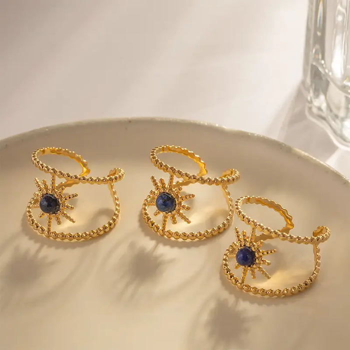 A collection of three gold open cuff bracelets, each featuring a central lapis lazuli gemstone within a detailed sunburst design, placed elegantly on a shallow ceramic dish under soft lighting. The glossy finish of the gold contrasts beautifully with the deep blue of the lapis lazuli, highlighting the intricate craftsmanship of the jewelry.