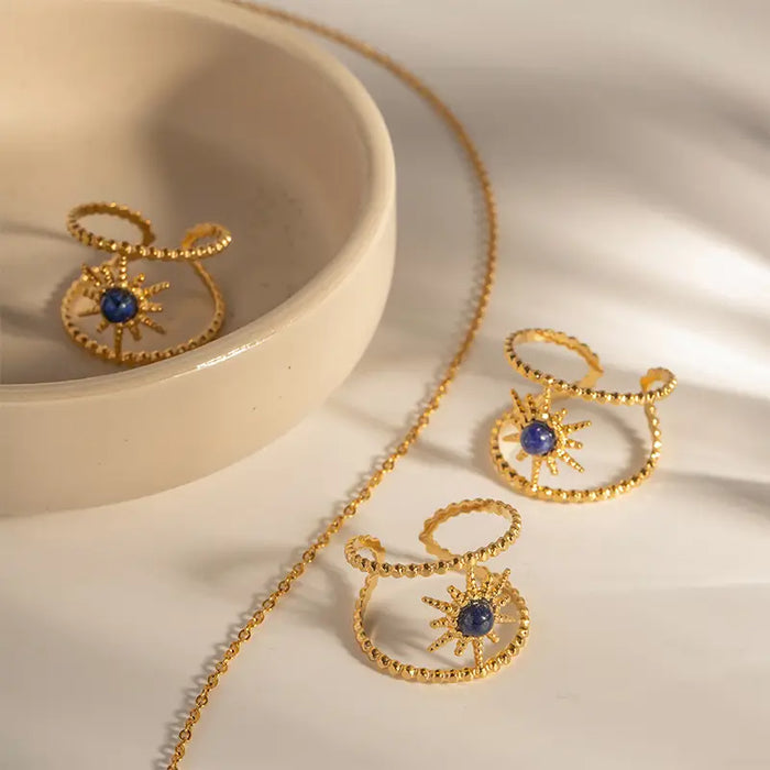 An elegant display of three lapis lazuli sunbeam bracelets on a smooth, shallow ceramic dish. Each gold bracelet features a radiant sunburst design with a prominent blue lapis lazuli stone at its center. The scene is bathed in warm, soft light, enhancing the rich gold tones and the deep blue of the gemstones, creating a luxurious and inviting presentation