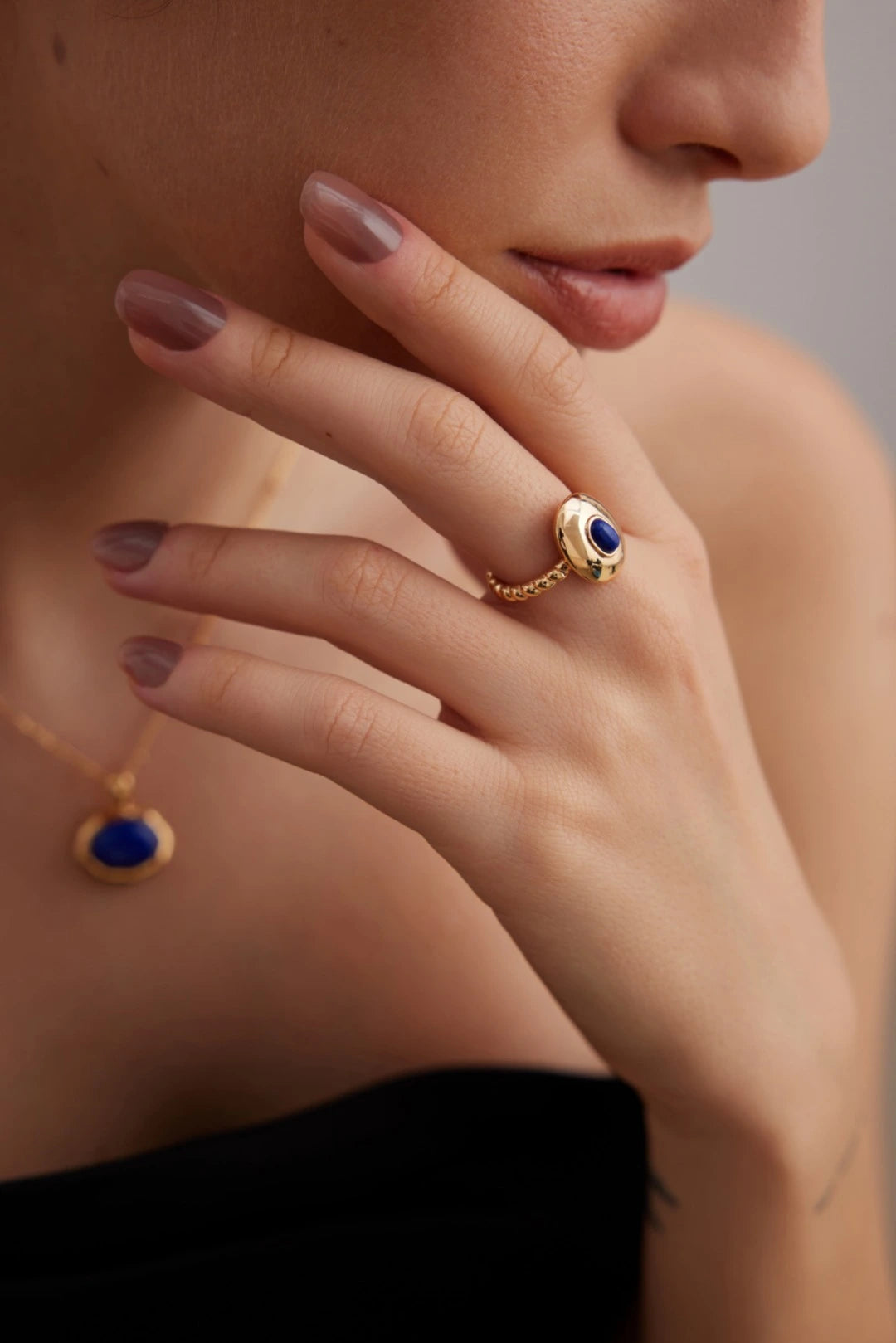 Close-up of a hand wearing a gold ring with a blue gemstone, matching necklace, on a neutral background.