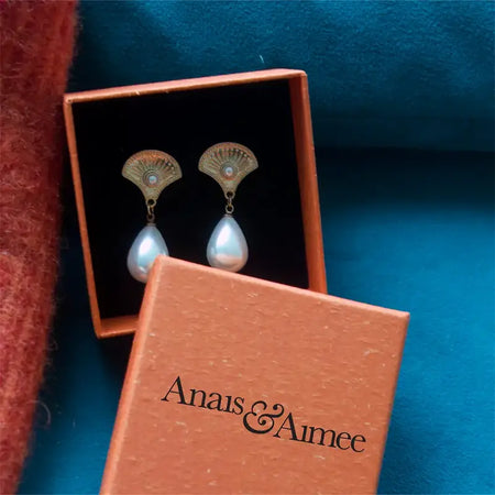 The image displays a pair of elegant earrings resting inside an open, terracotta-colored box with the brand name "Anais & Aimee" embossed in black on the box lid. The earrings feature a fan-shaped, ornately designed top in a gold-tone finish with a central gemstone, and a large, lustrous pearl drop hanging below. The background is a rich, blue fabric, enhancing the luxurious appearance of the earrings and the presentation box.