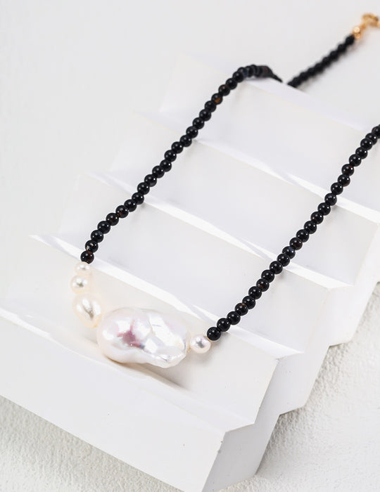 Elegant necklace featuring polished black onyx beads leading to a central large baroque pearl flanked by smaller pearls, displayed artistically on a white, tiered platform