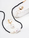 A sophisticated jewelry set displayed on a white surface, featuring a black onyx bead necklace with a large baroque pearl pendant, matched with gold earrings each holding a similar baroque pearl.