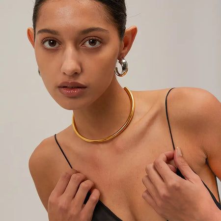  a close-up of a woman wearing unique and stylish jewelry. She has large, silver hoop earrings that have a reflective, polished finish, and a minimalist gold choker around her neck, enhancing her elegant look. Her bare shoulders and subtle makeup, combined with her direct gaze, give a sophisticate