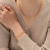 a woman dressed in a beige ribbed sweater, accessorized with a gold paperclip chain bracelet and necklace. The bracelet echoes the minimalist style of the necklace, both featuring elongated links that resemble paperclips. The jewelry set complements her outfit by adding a touch of elegance and refinement, highlighting her graceful hand pose and understated fashion choice. 