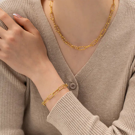 a woman dressed in a beige ribbed sweater, accessorized with a gold paperclip chain bracelet and necklace. The bracelet echoes the minimalist style of the necklace, both featuring elongated links that resemble paperclips. The jewelry set complements her outfit by adding a touch of elegance and refinement, highlighting her graceful hand pose and understated fashion choice. 