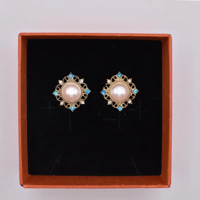 A pair of Royal Freshwater Pearl Stud Earrings displayed in an elegant box. Each earring features a central lustrous freshwater pearl surrounded by intricate gold detailing and accented with sparkling blue and white gemstones.