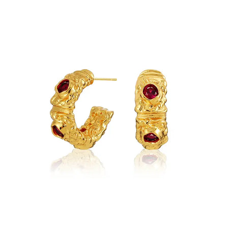 Close-up shot of a pair of gold hoop earrings with intricate designs and ruby gemstones against a white background.