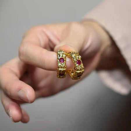 A hand holding a pair of gold gemstone hoop earrings, showing their rich texture and ruby detailing.