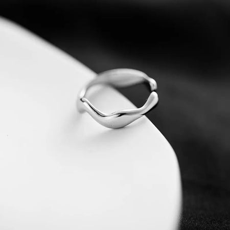 Close-up of a minimalistic silver cuff ring with a modern, twisted design, displayed against a blurred white background.