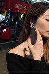 Woman wearing Serenity Black Onyx Silver Earrings from Anaïs & Aimee, standing in front of a classic red London double-decker bus at night. The bold, dark onyx stone contrasts with the surrounding polished silver, capturing a timeless and sophisticated style.